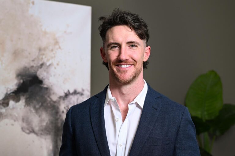 Headshot of Dr. Hildreth in a white dress shirt and dark blue blazer stands beside a modern wall adorned with abstract art featuring black and beige brushstrokes. A green plant adds a natural accent to the otherwise minimalist indoor setting.