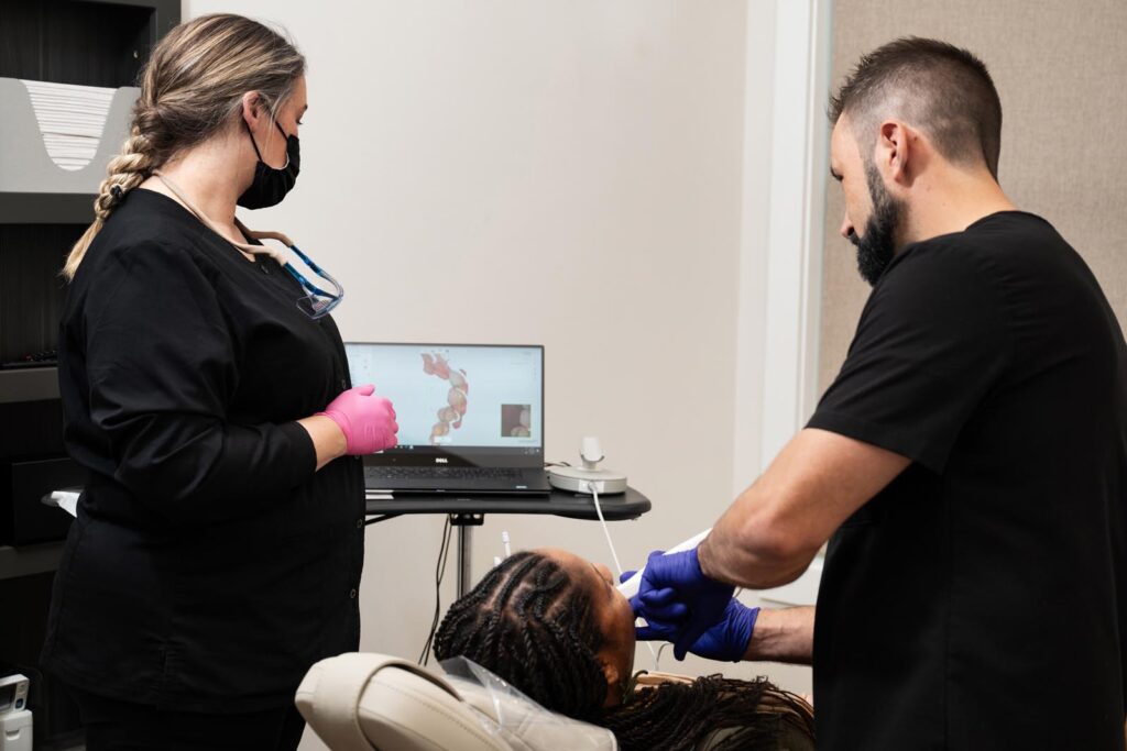 A dental professional in blue gloves performs a procedure on a reclining patient while a colleague in pink gloves observes. A laptop screen in the background displays a 3D dental model, indicating the use of advanced technology during the treatment.
