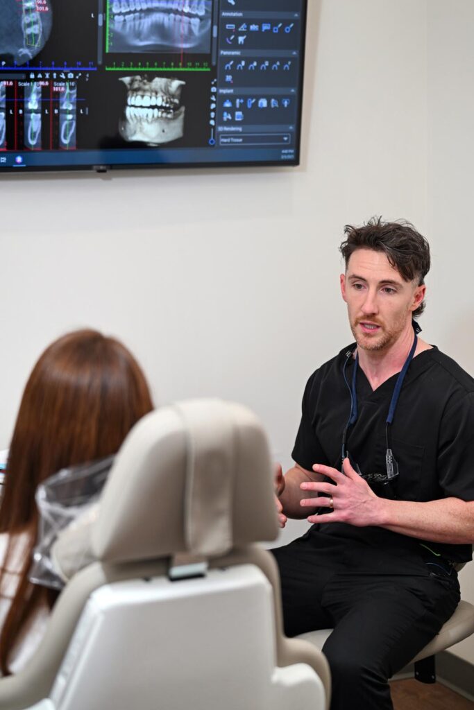 A dentist in black scrubs conducts a consultation with a female patient seated in a dental chair. Behind them, a wall-mounted screen displays detailed dental scans, including X-rays and 3D images, which appear to be actively referenced during the discussion.