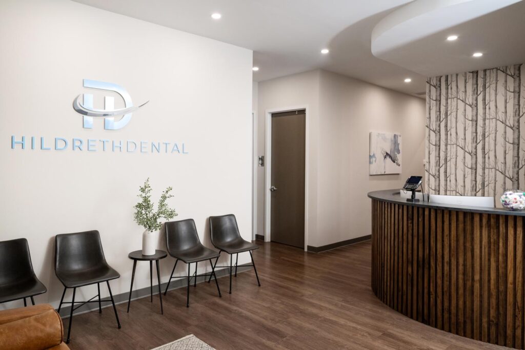 A sleek reception area at Hildreth Dental showcases a curved wooden desk with a computer, decorative bowl, and tree-patterned wallpaper behind it. To the side, four black chairs and a table with a plant create a cozy waiting space, framed by warm lighting and tasteful artwork.