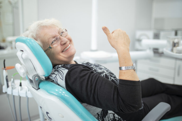 Older adult seated in a turquoise dental chair, smiling and giving a thumbs-up gesture while wearing glasses and a patterned top. The modern clinic setting includes visible dental equipment in the background, conveying a positive patient experience.