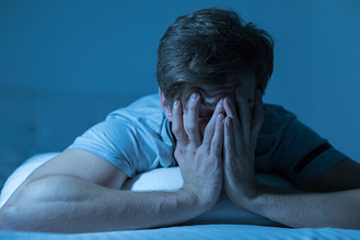 Person lying on bed in dim blue‑toned light with both hands covering face, conveying fatigue or stress.
